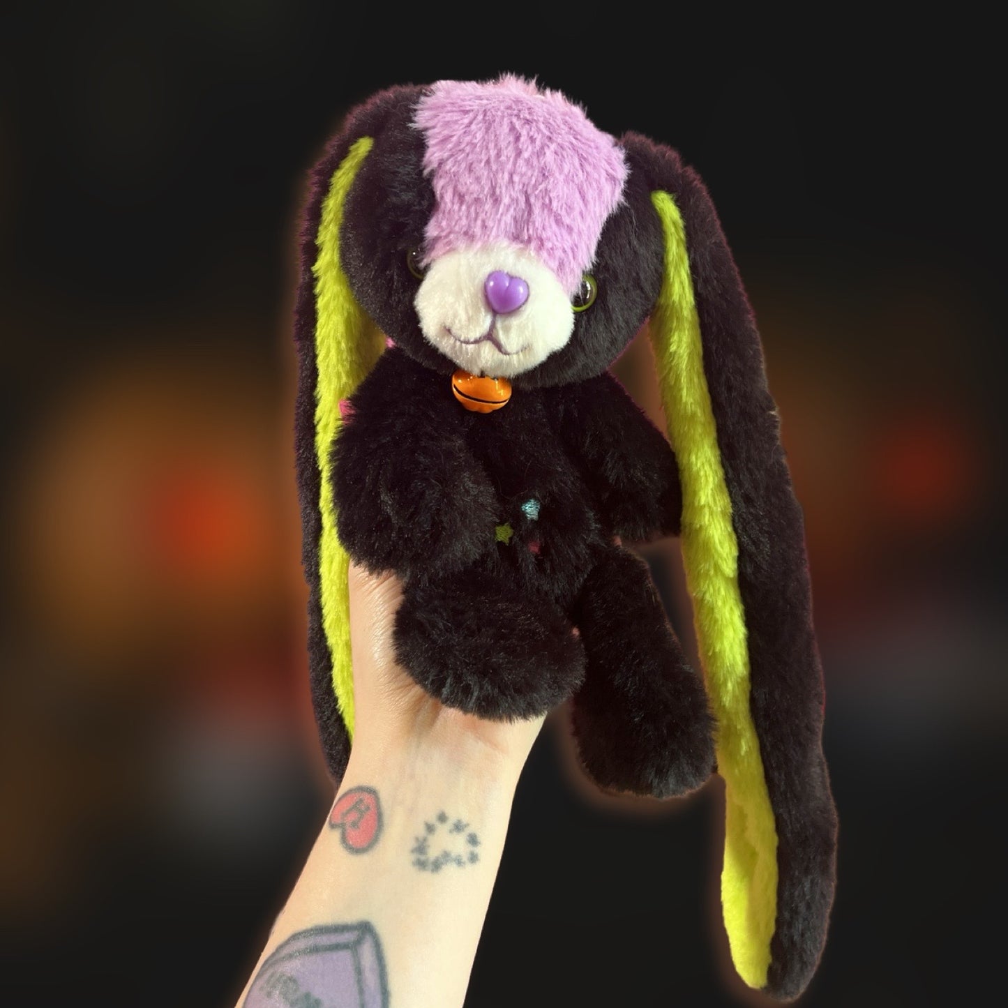 Plush doll bunny with a purple forehead stripe, black fur, and bright green ears. Plush has jack o lantern bell and is Halloween themed.